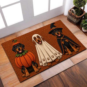 May include: A rectangular brown doormat featuring three dachshunds in Halloween costumes. The left dog wears a pumpkin suit, the center dog is draped in a white ghost sheet with paw prints, and the right dog wears a black witch hat and cape.