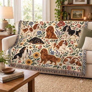 May include: A woven throw blanket featuring a cream background with a repeating pattern of Cavalier King Charles Spaniels in various colours, including black and tan, ruby, and blenheim, interspersed with colourful floral motifs and butterflies.
