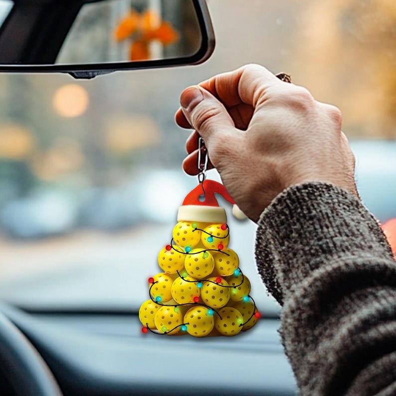 May include: A yellow and red Christmas tree bauble made of miniature yellow pickleball balls with red and green lights. The bauble is hanging from a rearview mirror in a car.