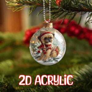 May include: A clear, round Christmas bauble featuring a small dog in a red and white outfit. The bauble hangs from a silver chain and has the words "2D ACRYLIC" in red and white. The background is blurred.