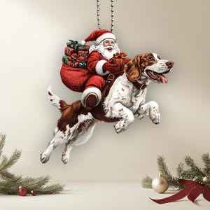 May include: A Christmas ornament featuring Santa Claus riding a brown and white dog with a red bag full of presents. The ornament is hanging from a silver chain.