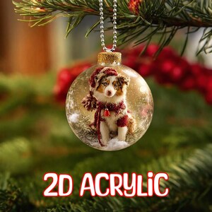 May include: A clear, round Christmas bauble featuring a dog in a red and white hat and scarf. The bauble hangs from a silver chain and has the words "2D ACRYLIC" in red and white at the bottom. The background is blurred.