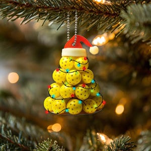 May include: A yellow pickleball Christmas tree bauble with a red Santa hat. The bauble is decorated with colourful lights.