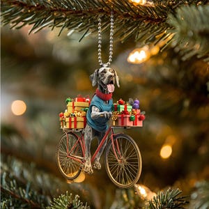 Puede incluir: Un adorno navideño con un perro montando una bicicleta con una cesta llena de regalos. El perro lleva un suéter azul y un collar rojo. La bicicleta es roja con una cesta negra y ruedas negras.