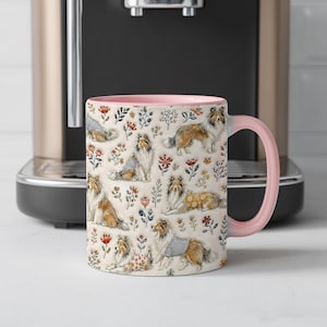 May include: A ceramic mug with a light pink interior and handle, featuring a repeating pattern of brown and white Shetland Sheepdogs interspersed with small red, orange, and blue floral illustrations on a cream-colored background.