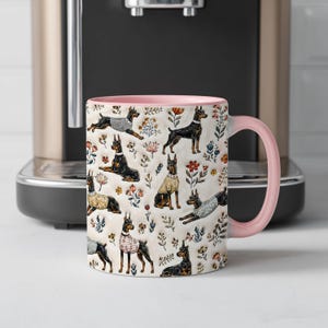 May include: A ceramic mug with a light pink handle and interior, featuring a repeating pattern of black and tan Doberman dogs wearing various sweaters, interspersed with small colorful floral illustrations on a cream-colored background.