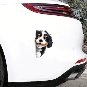 May include: A white car with a black and white dog sticker on the back bumper. The dog is peeking out from behind a white surface and has a happy expression.