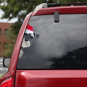 May include: A red car window sticker featuring a Schnauzer dog wearing a Santa hat. The dog's head and part of its body are visible, peeking out from behind the window. The sticker is designed to look like the dog is looking out of the car.