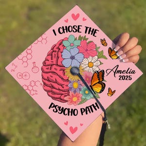 Op de afbeelding: Een roze afstudeerhoed met een ontwerp van een brein en bloemen. De tekst "I chose the psycho path" staat op de hoed gedrukt. De naam Amelia en het jaar 2025 staan ook op de hoed gedrukt.