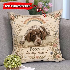 Op de afbeelding: Een vierkant sierkussen met een crèmekleurige achtergrond waarop een bruin-witte hond op een kussen ligt. Boven de hond staat een pastelkleurige regenboog en daaronder de tekst "Forever in my heart". Het ontwerp is omlijst door bloemranken met roze en blauwe bloemen.