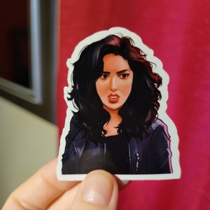 May include: A sticker featuring a digital illustration of a woman with dark hair and a leather jacket. She has a serious expression on her face.
