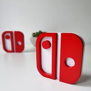 May include: Red and white 3D-printed decorative objects in the shape of a video game controller. The objects have a rectangular shape with a rounded edge and a circular button. The background is white.