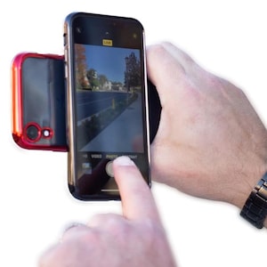 May include: A black smartphone with a red case attached to the back. The phone is in camera mode and the screen shows a live preview of a street scene. The word "LIVE" is displayed in the top right corner of the screen. The phone is being held by a hand, and a finger is pressing the "PHOTO" button on the screen.
