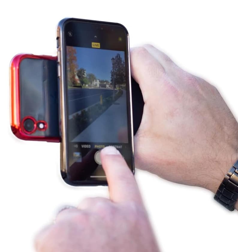 May include: A black smartphone with a red case attached to the back. The phone is in camera mode and the screen shows a live preview of a street scene. The word "LIVE" is displayed in the top right corner of the screen. The phone is being held by a hand, and a finger is pressing the "PHOTO" button on the screen.