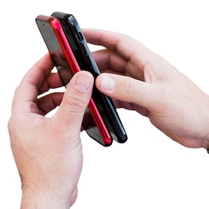 May include: Two smartphone cases side-by-side, one black and one red. The cases are held in a person's hands.