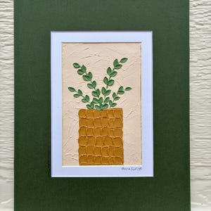 May include: An original painting of a plant in a square pot. The pot is mustard yellow, and the plant has green leaves. The painting is on a textured, light beige background, framed in white, and set against a dark green mat.