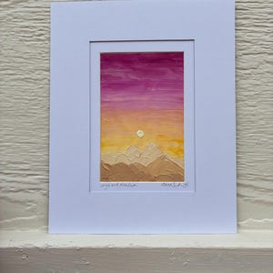 May include: An abstract landscape painting depicting a sunset over mountains. The sky transitions from pink to yellow, with a textured mountain range in shades of brown. The artwork is framed in white.