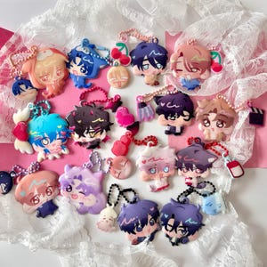 Puede incluir: Una colección de dijes acrílicos coloridos que presentan varios personajes de estilo anime con rostros expresivos, cada uno unido a una pequeña cadena de cuentas. Los dijes muestran diversos colores de cabello, incluyendo azul, morado, rosa y marrón.