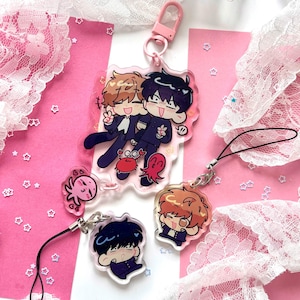 Puede incluir: Un conjunto de cuatro dijes de acrílico transparente con personajes estilo anime en uniformes escolares oscuros. El dije más grande incluye un broche de langosta rosa, mientras que los más pequeños tienen correas negras. Los diseños muestran dos personajes de cabello castaño y negro, acompañados por pequeñas ilustraciones de cangrejos y pulpos rojos.