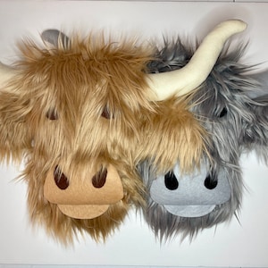 May include: Two plush Highland cow faces. One is tan with cream horns, the other is gray with gray horns. Both have felt features, including eyes, noses, and smiles. The plush has long, shaggy fur.