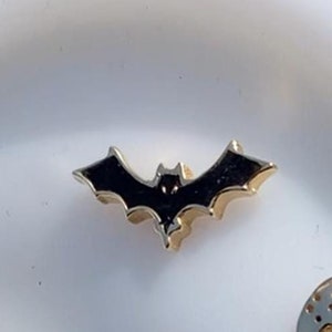 Könnte beinhalten: Nahaufnahme eines weißen Ohrmodells mit fünf goldenen Halloween-Ohrringen. Die Ohrringe zeigen eine Fledermaus, einen Schädel, ein Gespenst, einen Kürbis und eine Maske.