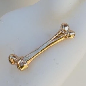Könnte beinhalten: Goldener Ohrring in Knochenform. Der Ohrring ist ein schlichtes Design mit abgerundeten Enden auf jeder Seite.
