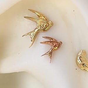 以下が含まれることがあります： 小さな鳥の形をしたジュエリーのクローズアップ。2つは焦点が合っており、1つは金、もう1つはローズゴールドで、翼と尾に詳細な特徴があります。他の作品はぼやけています。背景は柔らかいオフホワイトです。