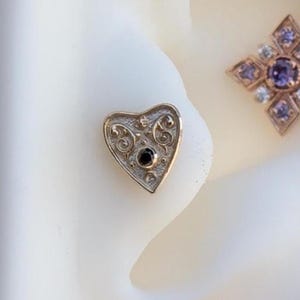 Puede incluir: Pendiente de corazón de oro con una piedra preciosa negra en el centro y detalles intrincados de filigrana.
