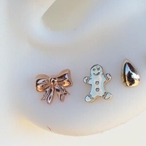 May include: Close-up of three small earrings: a rose gold bow, a silver gingerbread man, and a gold teardrop shape. The earrings are on a white surface. The bow earring has intricate details.