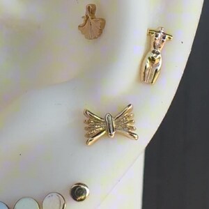May include: A close-up of a pair of gold earrings. The earrings are in the shape of a butterfly and a small human figure. The earrings are on a white background.