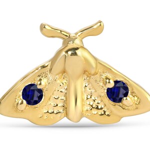 May include: A gold-colored moth-shaped jewelry piece with intricate detailing. The moth features two dark blue gemstones, one on each wing. The jewelry is set against a white background, highlighting the details of the design.