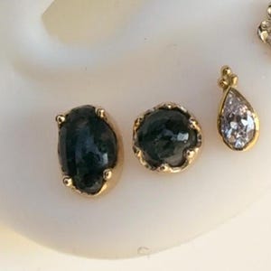 May include: Three gold-toned body jewellery pieces with green and clear gemstones. The two larger pieces are oval-shaped with green stones and gold accents. The smaller piece is a teardrop shape with a clear stone.