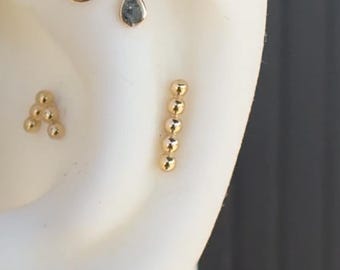 May include: A close-up of a person's ear with three gold earrings. The earrings are a small teardrop shape, a cluster of small balls, and a long bar with five small balls.