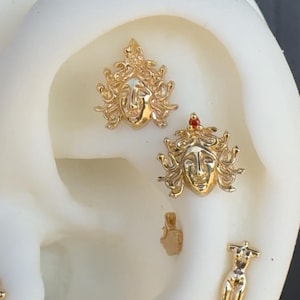 May include: Gold-toned earrings featuring a detailed Medusa head design. The earrings are shown on a mannequin's ear.