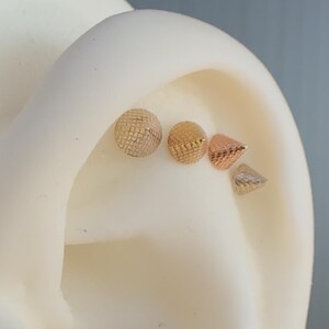 Puede incluir: Cuatro piercings de oreja dorados, dos son redondos y dos tienen forma de cono. Los piercings redondos tienen una superficie texturizada y los piercings con forma de cono tienen una superficie lisa.