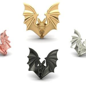 May include: A collection of bat-shaped jewelry pieces in various metallic finishes. The jewelry includes a rose gold stud, a gold pendant, a silver pendant, and a black pendant. The bats have detailed wings and bodies.