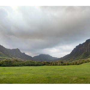 May include: A scenic landscape featuring a vibrant green field framed by towering mountains under a cloudy sky. The image captures a natural vista with lush vegetation and a dramatic, overcast atmosphere.