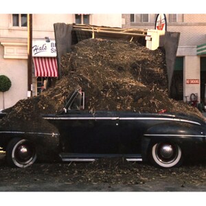 May include: A black vintage convertible car is overflowing with a large pile of dirt and debris. The car has white-walled tires and is parked in front of a building with a red and white striped awning. The scene appears to be outdoors.