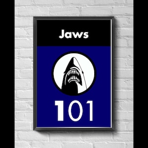 Może przedstawiać: Oprawiony plakat z czarną obwódką. Górna część jest czarna ze słowem "Jaws" w kolorze białym. Poniżej znajduje się niebieska sekcja z białym okręgiem zawierającym grafikę rekina. Numer "101" jest biały na dole.