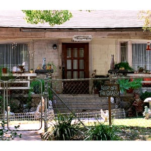 May include: A quaint, aged house with a wooden door and a sign that reads "Gartenrude Tannen". A "Keep Off The Grass" sign is visible. The house is surrounded by a chain-link fence and features decorative statues and potted plants.