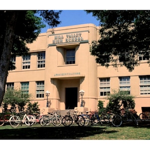 May include: Exterior view of Hill Valley High School, a tan-colored building with multiple windows and a central entrance. The words "Hill Valley High School" are displayed above the entrance. Bicycles are parked on the lawn in front of the building.