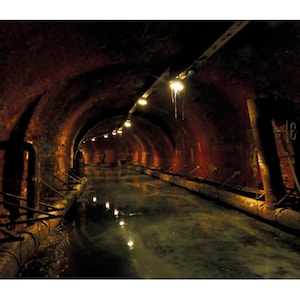 May include: An arched brick tunnel with a water-filled channel running through the center. Overhead, a series of lights illuminate the tunnel, casting reflections on the water. The tunnel's walls are a deep reddish-brown, creating a sense of depth and mystery.