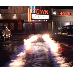 May include: Nighttime scene of a vintage movie theater with the marquee reading "TOWN" and "THE ATOMIC KID." Flames emanate from the road, with classic cars parked nearby. The scene is illuminated by neon lights.