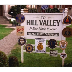 May include: A white sign that reads "Welcome to Hill Valley" with various circular and rectangular signs attached. The sign also says "A Nice Place To Live" and "Please Drive Carefully!"