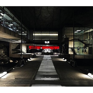 May include: An interior shot of a modern, industrial-style garage or workshop. A silver car is parked on a metal grate pathway, with a red-lit area in the background. The space features concrete walls, glass windows, and overhead lighting.