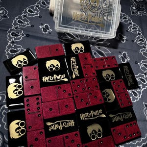 Op de afbeelding: Een set zwarte en rode glitterdominostenen met gouden Harry Potter-logo's en personagesilhouetten, vergezeld van een doorzichtige plastic opbergdoos met een gouden Harry Potter-afbeelding op het deksel, gerangschikt op een donkere stof.