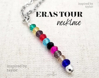 May include: A silver chain necklace with a beaded pendant featuring a rainbow of colors, inspired by the Eras Tour. The text "ERAS TOUR necklace" is written in black on a white background.