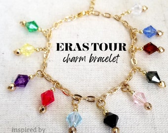May include: Gold chain bracelet with colorful crystal charms. The charms are in the shape of diamonds and are in various colors, including red, blue, green, yellow, purple, black, pink, and clear. The bracelet has the text "ERAS TOUR charm bracelet" on it.