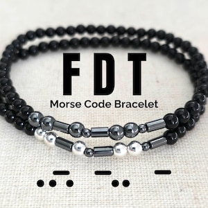May include: Black and silver beaded bracelet with the letters "FDT" and the words "Morse Code Bracelet". The bracelet is made with black beads and silver beads with silver bars in between. The Morse code for "FDT" is shown below the bracelet.