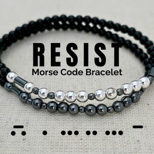 May include: A black and silver beaded bracelet with the word "RESIST" and the Morse code for the word. The bracelet is made of two strands of beads, one black and one silver.